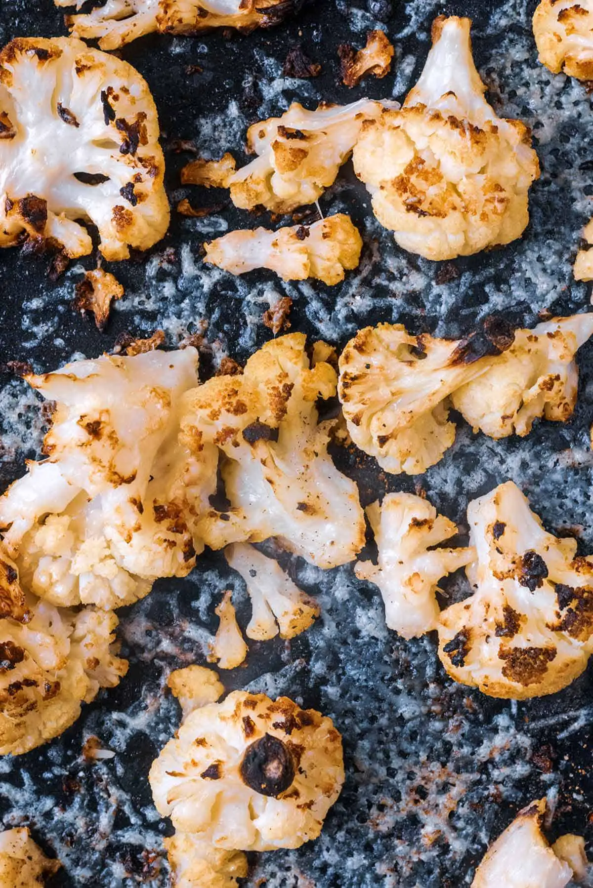 Cooked cauliflower florets and melted parmesan on a black baking tray,